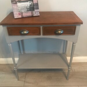 May include: A grey console table with two drawers and a lower shelf. The top of the table is a light brown wood. The table has turned legs and metal drawer pulls. A framed picture sits on the top of the table.