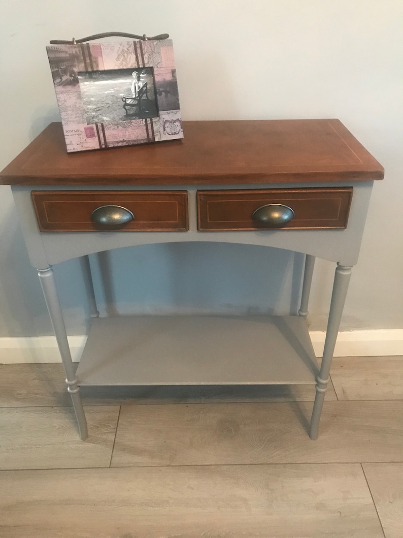 May include: A grey console table with two drawers and a lower shelf. The top of the table is a light brown wood. The table has turned legs and metal drawer pulls. A framed picture sits on the top of the table.
