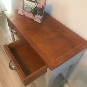 May include: A light blue wooden console table with a drawer and two shelves. The table has a distressed finish and a brown top. A picture frame with a pink and white design sits on the top of the table.