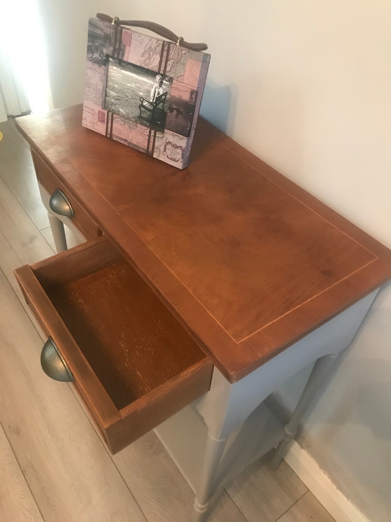 May include: A light blue wooden console table with a drawer and two shelves. The table has a distressed finish and a brown top. A picture frame with a pink and white design sits on the top of the table.