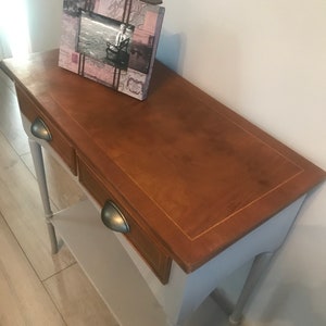 May include: A light grey console table with two drawers and a lower shelf. The top of the table is a darker wood colour with a light wood trim. The table has two turned legs and a picture frame on top.