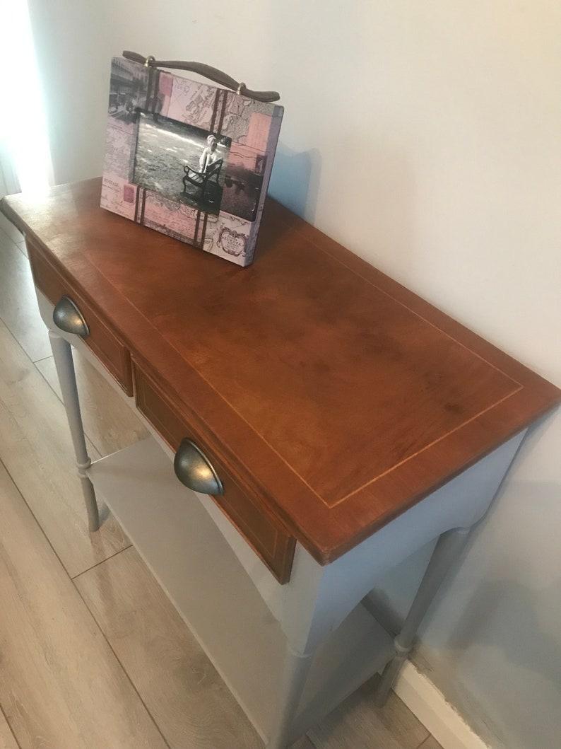 May include: A light grey console table with two drawers and a lower shelf. The top of the table is a darker wood colour with a light wood trim. The table has two turned legs and a picture frame on top.