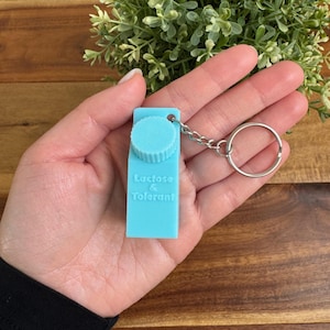 Op de afbeelding: Een lichtblauwe sleutelhanger in de vorm van een melkpak met een witte schroefdop. De sleutelhanger heeft de tekst "Lactose & Tolerant" op de voorkant gedrukt.