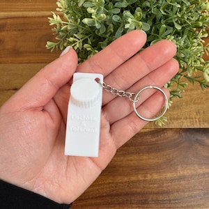 Op de afbeelding: Witte melkpak sleutelhanger met een zilveren ketting en een ring. Het pak heeft de tekst "Lactose & Tolerant" erop gedrukt.