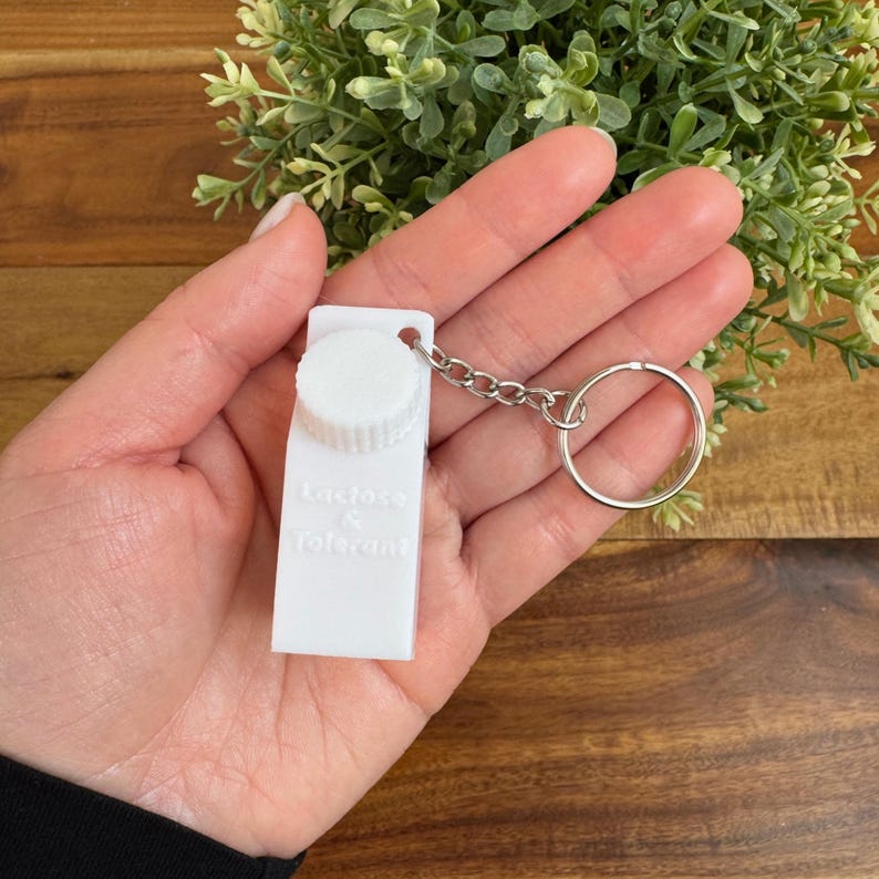 Op de afbeelding: Witte melkpak sleutelhanger met een zilveren ketting en een ring. Het pak heeft de tekst "Lactose & Tolerant" erop gedrukt.