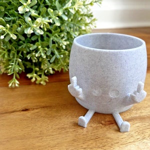 May include: A gray ceramic planter with a speckled finish, shaped like a cartoon character with arms and legs. The planter has two small circles for eyes and a small smile. The planter is sitting on a wooden surface.