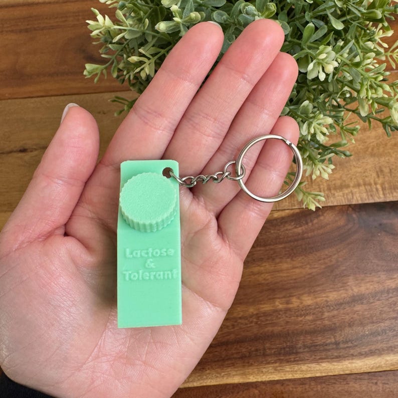 Op de afbeelding: Een groene sleutelhanger in de vorm van een melkpak met een wit deksel en de tekst "Lactose & Tolerant".