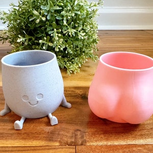May include: Two silicone cups with a fun design. One cup is light blue with a speckled finish and a smiling face. The other cup is pink and has a rounded shape.