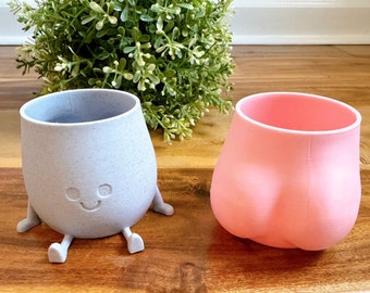 May include: Two silicone cups with a fun design. One cup is light blue with a speckled finish and a smiling face. The other cup is pink and has a rounded shape.