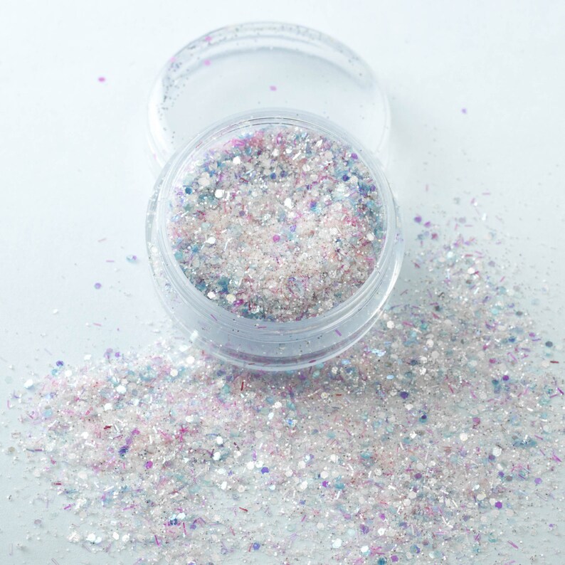 Puede incluir: Un tarro de pl&aacute;stico transparente lleno de purpurina iridiscente en tonos blanco, rosa, azul y morado. La tapa est&aacute; quitada y algo de purpurina est&aacute; esparcida alrededor. La purpurina es una mezcla de formas y tama&ntilde;os, creando un efecto brillante.