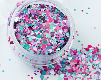 May include: A clear plastic jar filled with pink, teal, and iridescent glitter. The glitter is scattered on a white surface.