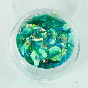 Peut inclure: Un récipient rond transparent rempli de paillettes vertes et bleues irisées. Les paillettes sont de différentes formes géométriques, reflétant la lumière. Le récipient est ouvert, révélant les paillettes à l'intérieur. L'effet général est scintillant et attrayant.
