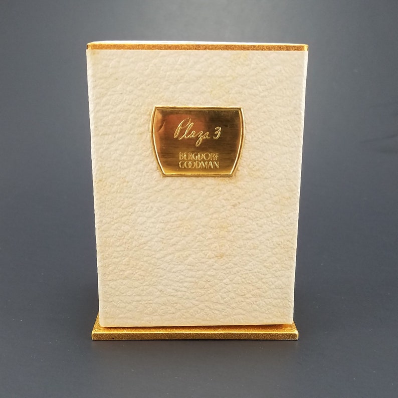 May include: A white box with a gold trim and a gold label that says "Plaza 3 Bergdorf Goodman".