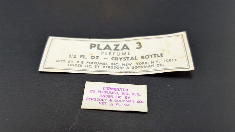May include: Two white labels with black text for Plaza 3 perfume. The top label reads "Plaza 3 Perfume 1/2 fl. oz. - Crystal Bottle Dist. by G Perfumes, Inc. New York, N.Y. 10012 Under Lic. by Bergdorf & Goodman Co.". The bottom label reads "Distributor BG Perfumes, Inc. N.Y. Under Lic. by Bergdorf & Goodman Co. Net 1/4 fl. oz."