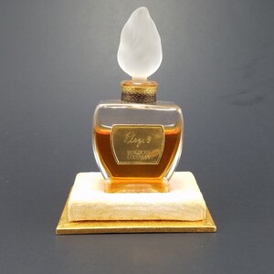 May include: A clear glass bottle of perfume with a gold cap and a frosted glass stopper shaped like a flame. The bottle is sitting on a small, square, cream-colored box with a gold trim. The label on the bottle reads "Claig's, Bergdorf Goodman".