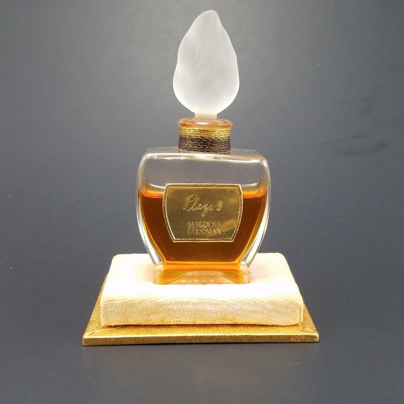 May include: A clear glass bottle of perfume with a gold cap and a frosted glass stopper shaped like a flame. The bottle is sitting on a small, square, cream-colored box with a gold trim. The label on the bottle reads "Claig's, Bergdorf Goodman".