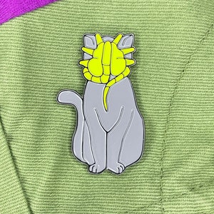 May include: A gray enamel pin of a cat with a yellow alien face. The cat is sitting with its paws together and its tail curled around its body. The alien face is a classic sci-fi design with a large head and eyes.