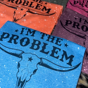 May include: A stack of colorful t-shirts with the text "I'M THE PROBLEM" and a bull skull graphic. The shirts are in various colors, including orange, blue, purple, and gray, with a speckled design.