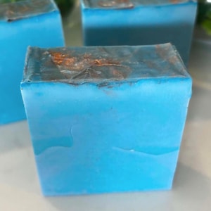 May include: A rectangular bar of soap with a vibrant blue body and a textured, metallic copper-colored top edge. The soap has a smooth, solid appearance on the front face, contrasting with the uneven, shimmering finish along the top surface.