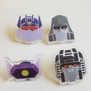 May include: Four acrylic pins featuring stylized character head designs. The top left is purple and blue, the top right is grey and black, the bottom left is a purple hexagon with a yellow center, and the bottom right is black and grey with red eyes.