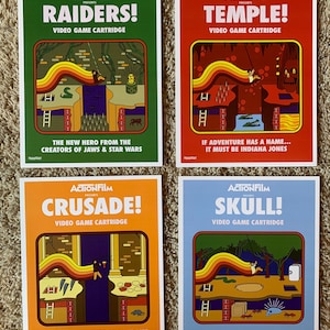 May include: Four retro-style video game cartridge covers featuring illustrations of Indiana Jones-themed adventures. The covers are green, red, orange, and blue with the titles "RAIDERS!", "TEMPLE!", "CRUSADE!", and "SKULL!" printed in white.