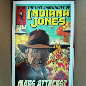 May include: Comic book cover for "The Lost Adventures of Indiana Jones" featuring a portrait of Indiana Jones, the text "Mars Attacks?", and illustrations of aliens and explosions. The cover has a green, yellow, and red color scheme.