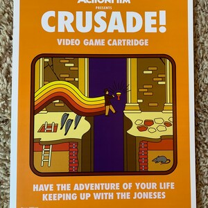 May include: A retro-style video game cartridge poster with an orange background. The poster features the text "ACTIONFILM PRESENTS CRUSADE! VIDEO GAME CARTRIDGE" and an illustration of a character holding a chalice, standing on a rainbow-colored path, with a ladder and a mouse in the background. The text "HAVE THE ADVENTURE OF YOUR LIFE KEEPING UP WITH THE JONESES" is at the bottom of the poster.