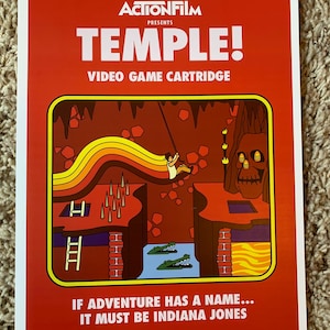 May include: A retro-style video game cartridge cover with a red background and the text "ACTIONFILM PRESENTS TEMPLE! VIDEO GAME CARTRIDGE". The cover features an illustration of a person swinging on a rope over a pit of spikes, crocodiles, and a skull. The text "IF ADVENTURE HAS A NAME... IT MUST BE INDIANA JONES" is at the bottom of the cover.