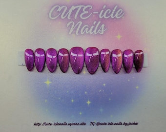 May include: A set of ten oval-shaped, press-on nails in a gradient of purple hues, with a white bow-like design at the base. The nails are displayed on a white strip against a background with the text "CUTE-icle Nails".