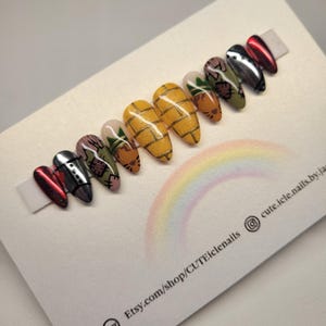 May include: A set of almond-shaped false nails with various designs. The nails feature a pineapple pattern, floral designs, and metallic finishes in red, silver, and black. The nails are displayed on a white surface with a rainbow and text.