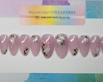 May include: Set of oval-shaped, light pink press-on nails. Each nail features a silver foil design with green and gold leaf accents. The nails are displayed on a white card with a sticker that includes a website and Instagram handle.