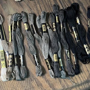 May include: A collection of DMC embroidery floss skeins arranged in a gradient from light gray to black. Each skein features a gold and black paper label with specific color numbers printed on them, including 645, 646, 844, and 310.