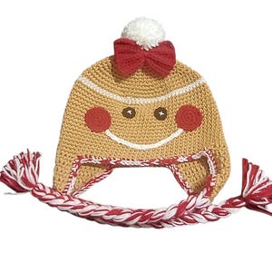 May include: A knitted gingerbread man hat with a smiling face, red cheeks, and a red bow. The hat has a white pom-pom and braided earflaps with red and white yarn. The hat is designed for children and is perfect for the holiday season.