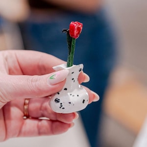 May include: A small white ceramic vase with black speckles, holding a single red flower with a green stem.