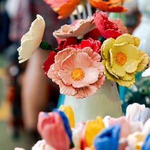May include: A vibrant display of artificial flowers in a pale green vase. The arrangement features red, orange, yellow, and pink flowers, including poppies and tulips. The flowers are arranged in a decorative bouquet.