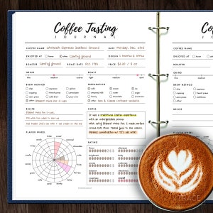 May include: A coffee tasting journal page featuring a brown and white coffee cup with a heart design in the foam. The page includes sections for coffee name, origin, roast date, grind, brew method, preparation, recipe, notes, and a flavour wheel.