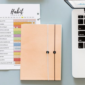Puede incluir: Un rastreador de h&aacute;bitos imprimible con una cuadr&iacute;cula colorida y una lista de h&aacute;bitos para rastrear, como levantarse temprano, preparar el desayuno y leer un libro. El rastreador est&aacute; impreso en papel blanco y est&aacute; al lado de una libreta de color melocot&oacute;n con un cierre el&aacute;stico blanco y negro.