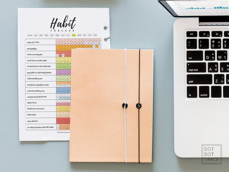 Puede incluir: Un rastreador de h&aacute;bitos imprimible con una cuadr&iacute;cula colorida y una lista de h&aacute;bitos para rastrear, como levantarse temprano, preparar el desayuno y leer un libro. El rastreador est&aacute; impreso en papel blanco y est&aacute; al lado de una libreta de color melocot&oacute;n con un cierre el&aacute;stico blanco y negro.