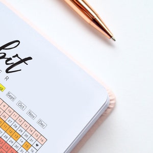Pode incluir: Um rastreador de h&aacute;bitos com uma capa rosa e uma caneta dourada. O rastreador tem um layout de calend&aacute;rio com os meses de junho a dezembro. A palavra "Habit Tracker" est&aacute; escrita em preto no topo da p&aacute;gina.