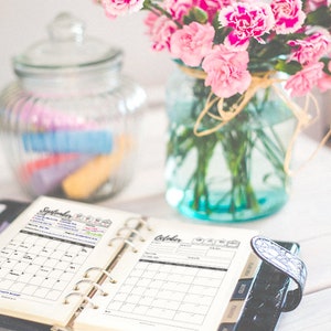 May include: An open planner with a black cover and silver rings. The planner has two open pages, one with a calendar for September and the other with a calendar for October. The planner is on a white wooden table with a glass jar of colorful items and a vase of pink carnations in the background.