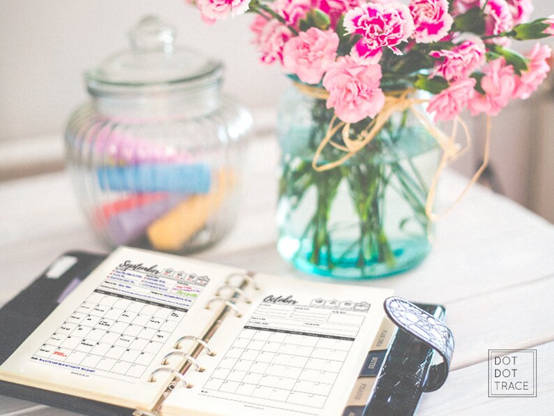 May include: An open planner with a black cover and silver rings. The planner has two open pages, one with a calendar for September and the other with a calendar for October. The planner is on a white wooden table with a glass jar of colorful items and a vase of pink carnations in the background.