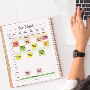 May include: A white planner with a weekly class schedule. The planner has a spiral binding and is open to the week of the month. The planner is on a white table with a laptop and a person's hand on the keyboard.