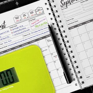 May include: A close-up of a yellow digital scale with a black and white calendar open to August and September. The scale reads 0.00 and the calendar has checkboxes for tasks and goals.