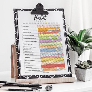 Pode incluir: Um rastreador de h&aacute;bitos com uma grade colorida e uma lista de h&aacute;bitos para rastrear, como "Beba &aacute;gua", "Medite" e "Leia algo novo". O rastreador est&aacute; em uma prancheta com um padr&atilde;o preto e branco com um suporte de madeira. A imagem tamb&eacute;m inclui uma planta verde em um vaso branco e uma pequena suculenta em um vaso cinza.