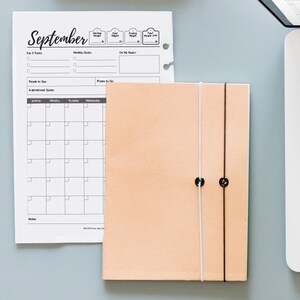 May include: A peach-colored leather bound journal with two white and black strings holding it closed. The journal is sitting on a gray surface next to a laptop computer and a monthly calendar planner with the word "September" at the top.