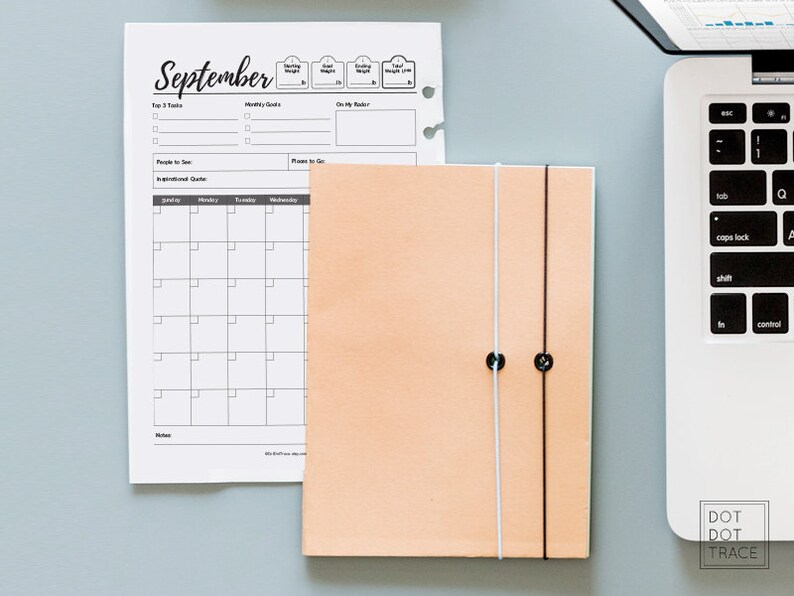 May include: A peach-colored leather bound journal with two white and black strings holding it closed. The journal is sitting on a gray surface next to a laptop computer and a monthly calendar planner with the word "September" at the top.