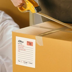 May include: A brown cardboard box with a white label that says "Kitchen" and has a list of items. The box is being taped shut with a yellow tape dispenser. The label also has a box that says "Box Contents" and a list of items including "Pottery Flower Set", "Blue Cups (18EA)", "Tripod Market (GREY)", and "Water". The label also has a section for "Floor", "Fragile", and "Heavy". The box is labelled with the number "10".