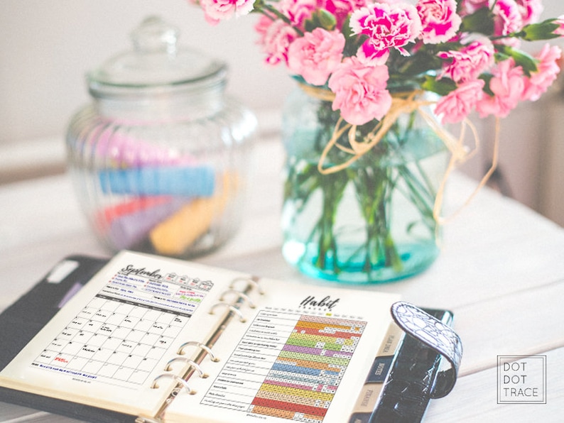 Puede incluir: Una carpeta negra con un calendario y p&aacute;ginas de seguimiento de h&aacute;bitos abiertas. La p&aacute;gina del calendario muestra el mes de septiembre. La p&aacute;gina de seguimiento de h&aacute;bitos tiene una cuadr&iacute;cula colorida con diferentes secciones para realizar un seguimiento de los h&aacute;bitos. La carpeta est&aacute; sobre una mesa de madera blanca con un frasco de vidrio de marcadores coloridos y un jarr&oacute;n de flores rosas.