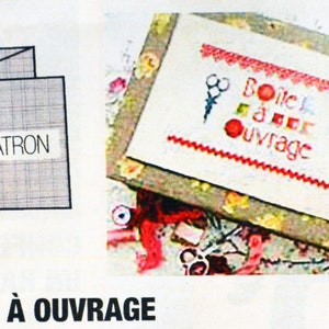 Könnte beinhalten: Eine weiße Stoffbox mit einem roten und weißen Stickmuster, auf dem "Boite a ouvrage" mit einer Schere und einer roten Kugel steht. Die Box ist von einem Blumenmuster umgeben.