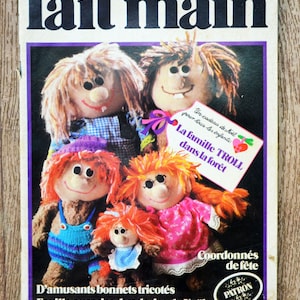 May include: A magazine cover for a craft publication called "fait main" with the text "Pas à Pas" and "Novembre 1982 Mensuel". The cover features a family of four knitted troll dolls with colorful clothing and a red heart-shaped tag that reads "Un cadeau de Noël pour tous les enfants: La famille TROLL dans la forêt". The text "Coordonnés de fête" and "D'amusants bonnets tricotés Feuille-surprise: broderies de Noël" is also visible on the cover.